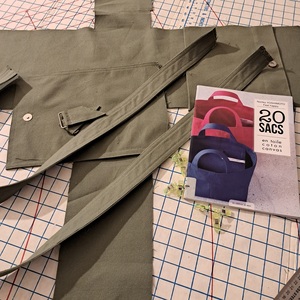 Olive green canvas cut out in the shape of a bag pattern resting on a cutting board, with other finished bag pieces (handle, straps, and flap), a book of bag patterns, and a bunch of wonderclips.