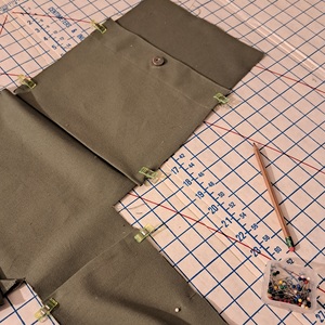 An image showing the front and lateral pockets pinned in place on an olive green bag body, resting on a cutting board. A pencil and container of pins are also visible.