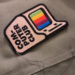 A close-up showing a patch with a computer and the words 'Computer Club' being stitched onto olive green canvas.